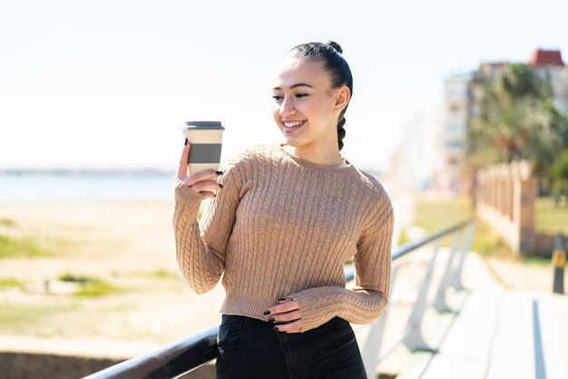 幸せな表情で屋外でテイクアウト コーヒーを保持している若いモロッコの女の子