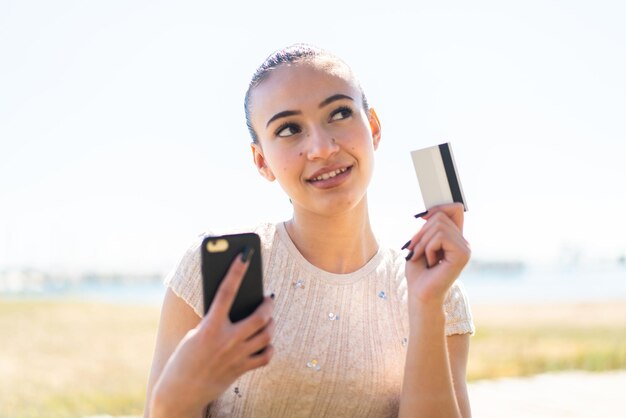 Young moroccan girl at outdoors buying with the mobile with a credit card while thinking