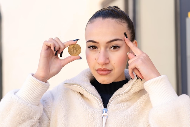 Young moroccan girl at outdoors holding a Bitcoin and having doubts