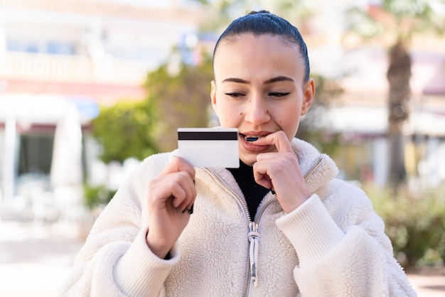 Young moroccan girl at outdoors holding a credit card and having doubts