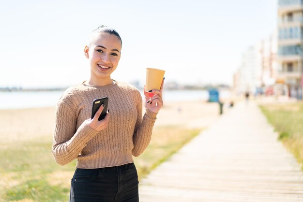 屋外で携帯電話を使用し、幸せそうな表情でコーヒーを保持している若いモロッコの女の子