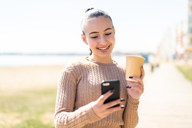 young moroccan girl at outdoors using mobile phone and holding a coffee with happy expression