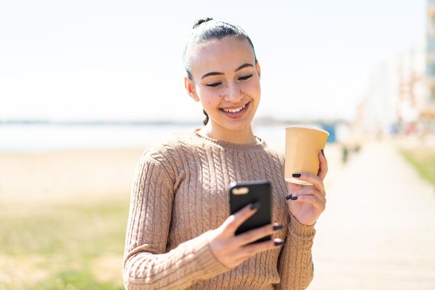 Young moroccan girl at outdoors using mobile phone and holding a coffee with happy expression