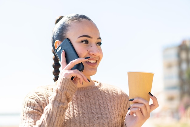 Giovane ragazza marocchina all'aperto utilizzando il telefono cellulare e tenendo un caffè con un'espressione felice