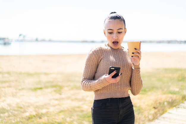 Young moroccan girl at outdoors using mobile phone and holding a coffee with surprised expression