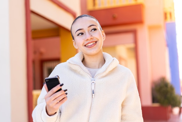 Young moroccan girl using mobile phone at outdoors looking up while smiling