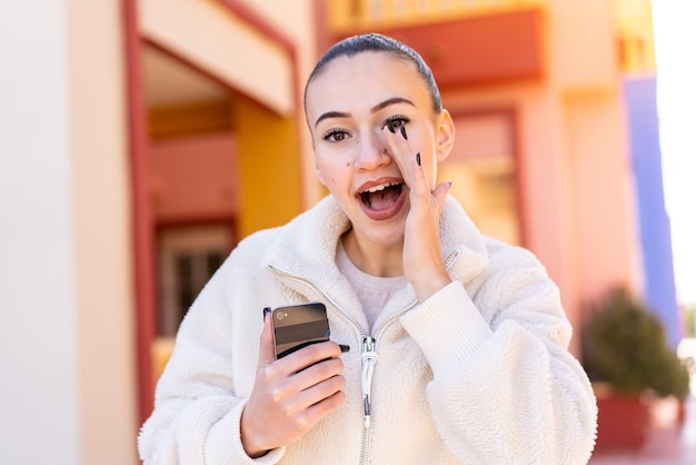 Young moroccan girl using mobile phone at outdoors shouting with mouth wide open