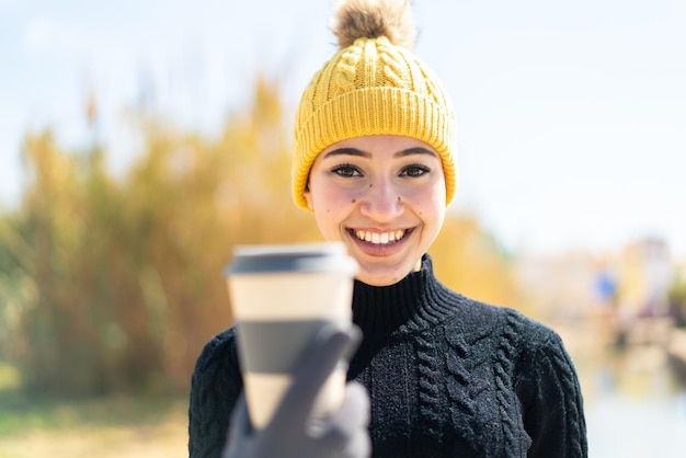 Молодая марокканская девушка в зимних муфтах, держащая кофе на улице со счастливым выражением лица