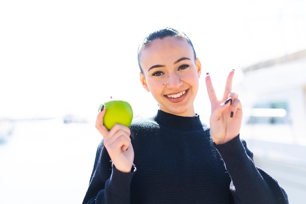 写真 屋外笑顔で勝利のサインを示すリンゴを持つ若いモロッコの女の子