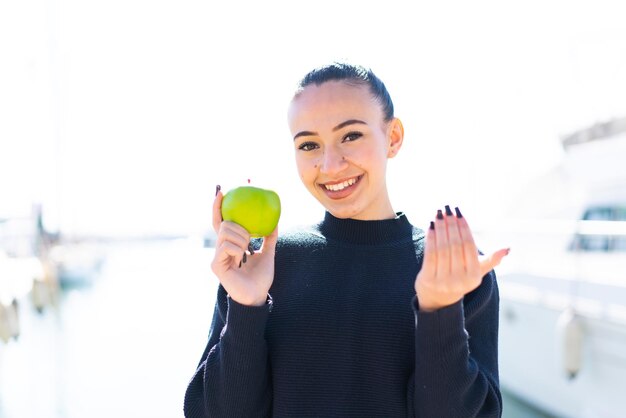 Photo young moroccan girl with an apple at outdoors inviting to come with hand happy that you came