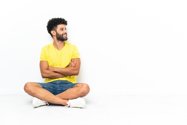 Foto giovane uomo bello marocchino seduto sul pavimento su sfondo isolato felice e sorridente