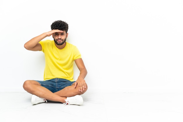 Young Moroccan handsome man sitting on the floor over isolated background looking far away with hand to look something