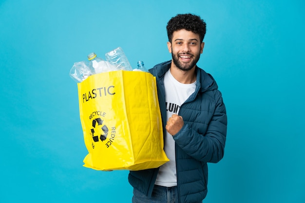 Young Moroccan man holding a bag full of plastic bottles to recycle over isolated wall celebrating a victory