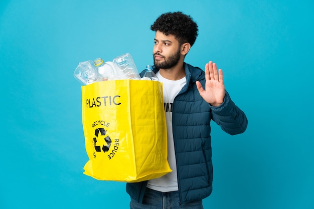 Foto giovane uomo marocchino che tiene un sacchetto pieno di bottiglie di plastica da riciclare facendo gesto di arresto e deluso