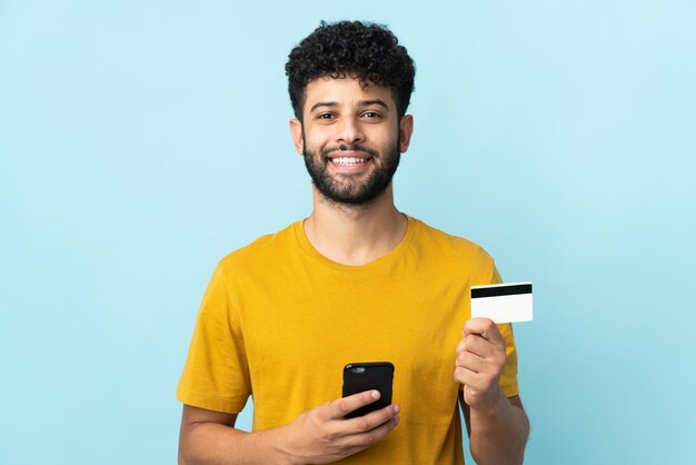 Young Moroccan man isolated on blue buying with the mobile with a credit card