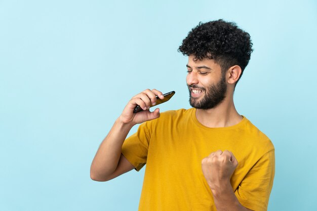 Young Moroccan man isolated on blue using mobile phone and singing