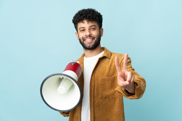 Young Moroccan man isolated on blue wall holding a megaphone and smiling and showing victory sign