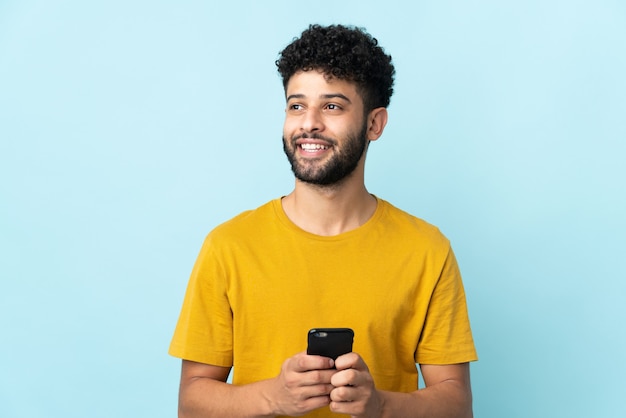 Young Moroccan man isolated on blue wall using mobile phone and looking up