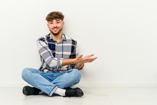 手のひらにコピースペースを保持している白で隔離の床に座っている若いモロッコ人男性。