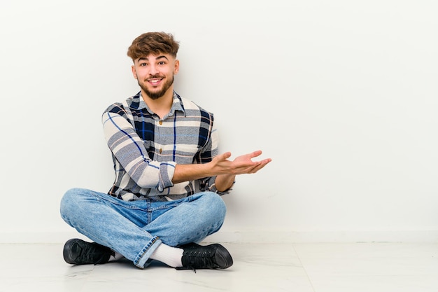 Young Moroccan man sitting on the floor isolated on white holding a copy space on a palm.