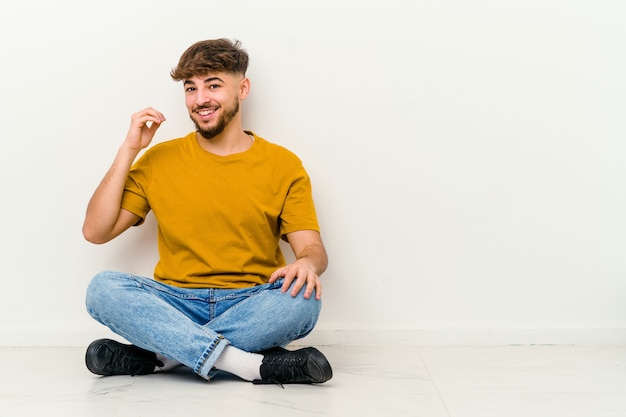 Foto giovane uomo marocchino seduto sul pavimento isolato su bianco che ride di qualcosa, che copre la bocca con le mani.