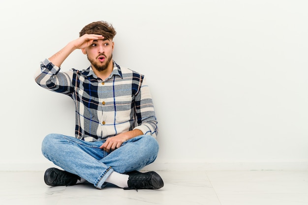 Young Moroccan man sitting on the floor isolated on white looking far away keeping hand on forehead.