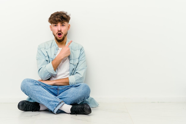 Young Moroccan man sitting on the floor isolated on white pointing to the side