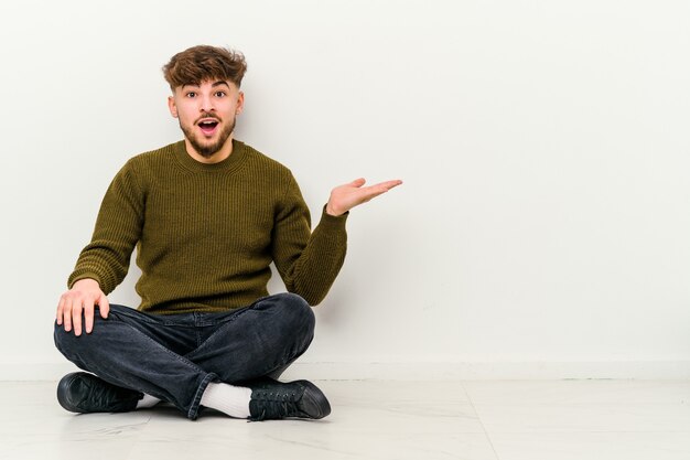 Young Moroccan man sitting on the floor isolated on white wall impressed holding copy space on palm.