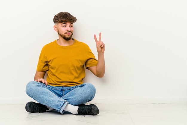 白い壁に隔離された床に座っている若いモロッコ人は、指で平和のシンボルを喜んで気楽に示しています。