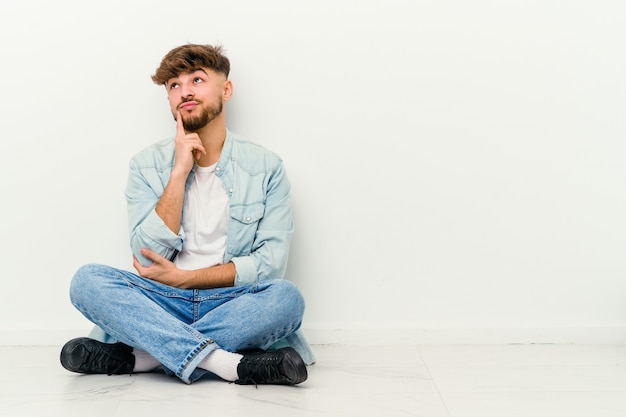 젊은 모로코 남자는 의심스럽고 회의적인 표정으로 옆으로보고 흰 벽에 고립 된 바닥에 앉아.