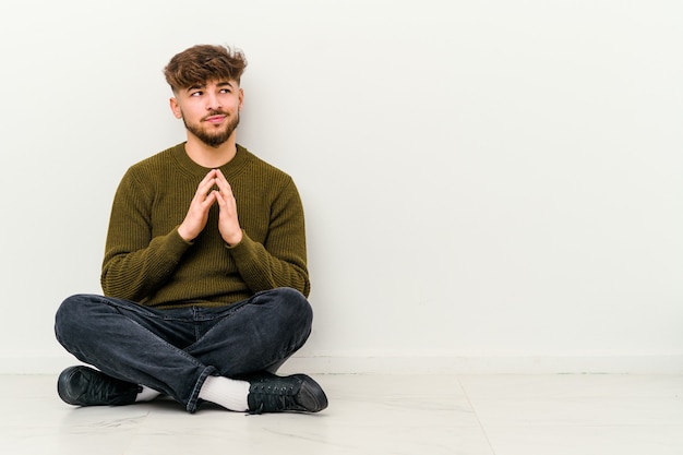 Young Moroccan man sitting on the floor isolated on white wall making up plan in mind, setting up an idea.