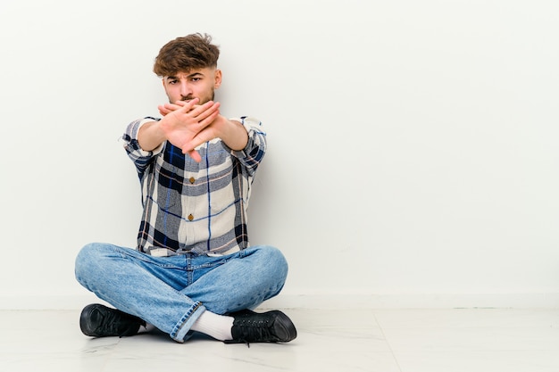 Young Moroccan man sitting on the floor isolated on white wall standing with outstretched hand showing stop sign, preventing you.