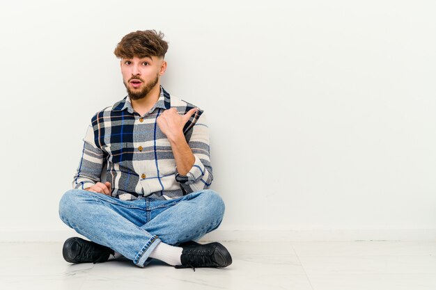 Young Moroccan man sitting on the floor   points with thumb finger away, laughing and carefree.