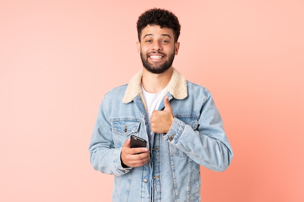 Young Moroccan man using mobile phone isolated on pink background giving a thumbs up gesture
