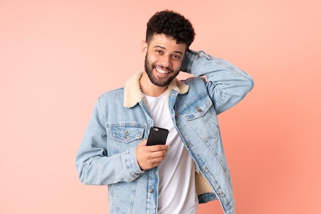 Young Moroccan man using mobile phone isolated on pink background laughing