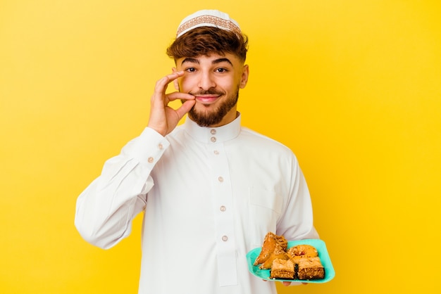 Young Moroccan man wearing the typical arabic costume eating Arabian sweets isolated on yellow background with fingers on lips keeping a secret.