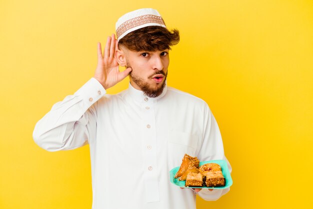 Giovane uomo marocchino che indossa il costume arabo tipico che mangia i dolci arabi isolati sulla parete gialla cercando di ascoltare un pettegolezzo.