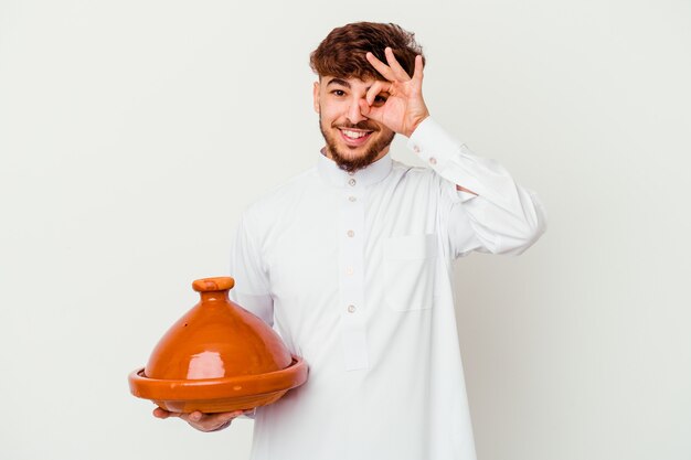 Young Moroccan man wearing the typical arabic costume holding a tajine isolated on white excited keeping ok gesture on eye.