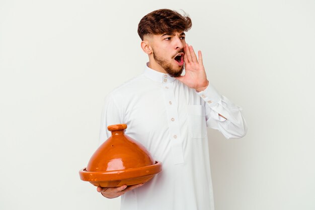 Young Moroccan man wearing the typical arabic costume holding a tajine isolated on white shouting and holding palm near opened mouth.