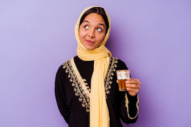 Young Moroccan woman holding a glass of tea isolated on purple background dreaming of achieving goals and purposes