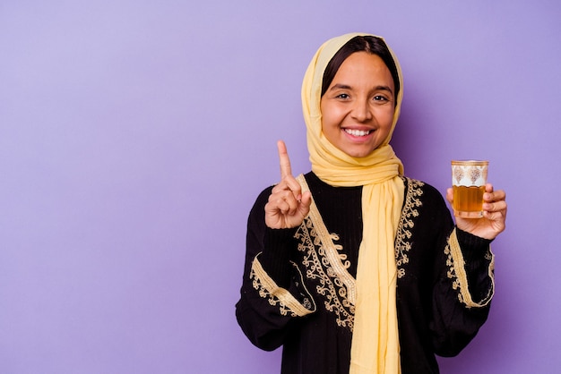 Giovane donna marocchina con in mano un bicchiere di tè isolato su sfondo viola che mostra il numero uno con il dito.