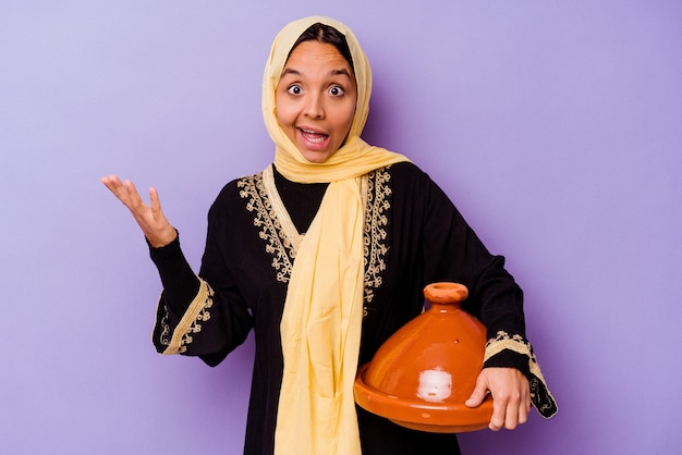 紫色の背景に分離されたタジンを持っている若いモロッコ人女性は、嬉しい驚きを受け取り、興奮し、手を上げます。