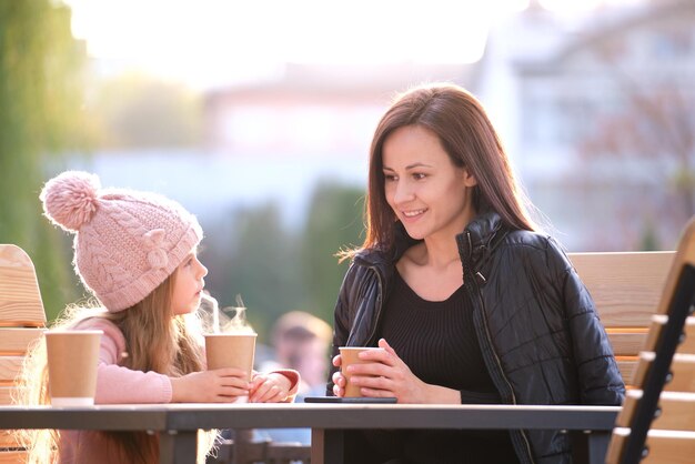 写真 晴れた秋の日にホットドリンクと一緒にストリートカフェに座って楽しい時間を過ごしている若い母と彼女の子供の娘家族関係の概念の幸せ