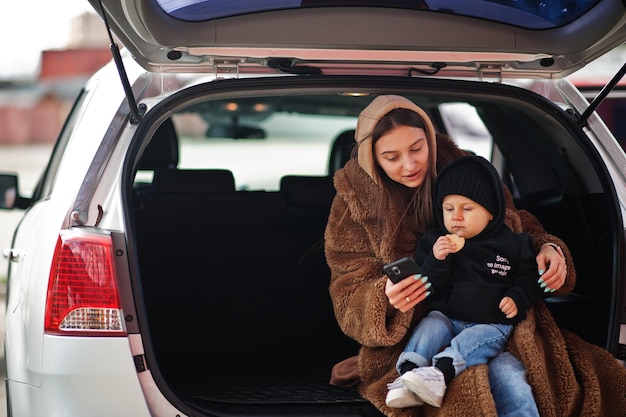 Young mother and child sitting in the trunk of a car and looking at mobile phone Safety driving concept