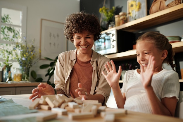 Young mother and cute daughter laughing at destroyed tower from wooden blocks