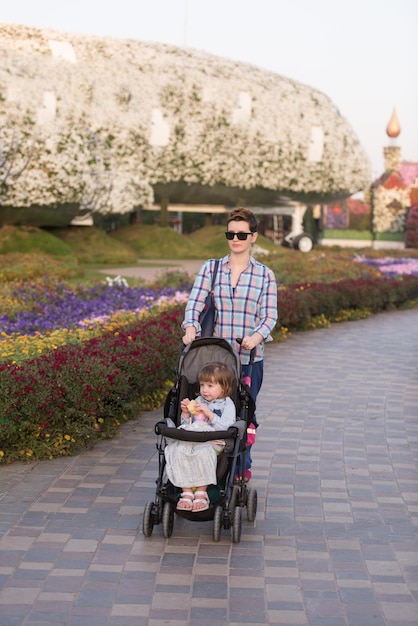 young mother and cute little girl in a baby stroller enjoying a beautiful day in the flower garden