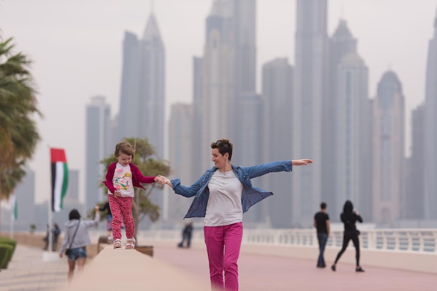 young mother and cute little girl running and cheerfully spend their time on the promenade by the sea with a big city in the background