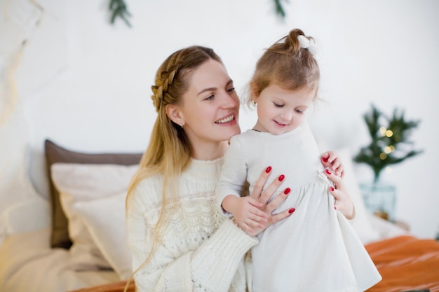 Young mother and daughter near Christmas tree Happy family waiting for Christmas and New year Cozy light interior with Christmas decorations and lights