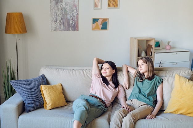 Молодая мать и дочь отдыхают на диване в гостиной у стены с задышками, лампой, зелеными домашними растениями и некоторой мебелью