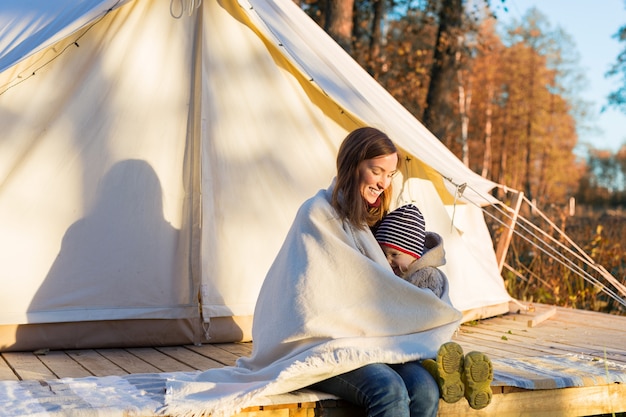 Young mother embracing her little kid with a blanket while sitting near canvas bell tent